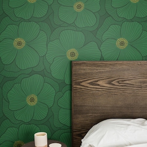 May include: Wallpaper featuring a repeating pattern of large green flowers with dark green outlines and yellow centers. The design is displayed on a wall behind a dark wood headboard, a white pillow, and a small round wooden side table with a candle.