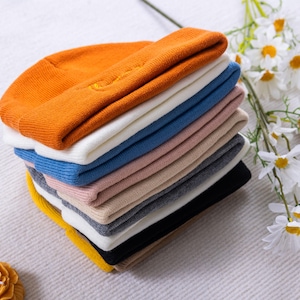 May include: A stack of colorful knit beanies in various colors, including orange, white, blue, pink, and black. The beanies are neatly folded and stacked on top of each other. A sprig of white daisies is in the background.