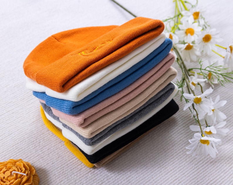 May include: A stack of colorful knit beanies in various colors, including orange, white, blue, pink, and black. The beanies are neatly folded and stacked on top of each other. A sprig of white daisies is in the background.