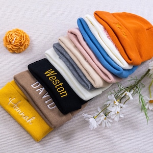 May include: A collection of colorful beanies in various shades, including yellow, brown, black, gray, pink, blue, white, and orange. Some beanies have embroidered names in white or yellow. A yellow flower-shaped candle and white flowers with green stems are also present.