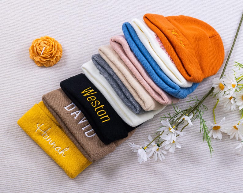 May include: A collection of colorful beanies in various shades, including yellow, brown, black, gray, pink, blue, white, and orange. Some beanies have embroidered names in white or yellow. A yellow flower-shaped candle and white flowers with green stems are also present.