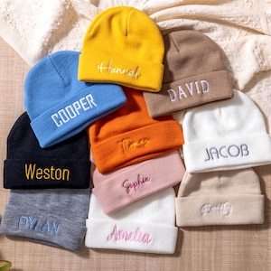 May include: A selection of colourful beanies, each embroidered with a name. The beanies are in shades of blue, yellow, orange, and white. Names include "Hannah", "Cooper", "Weston", "David", "Jacob", "Sophie", "Amelia", "Dylan", and "Family".