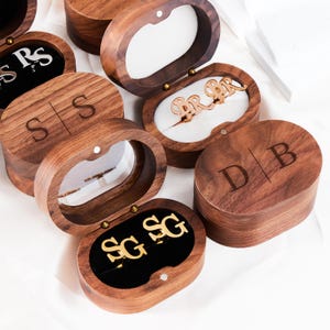 May include: Several wooden jewelry boxes with personalized initials are displayed. The boxes are oval-shaped and made of dark wood. Some boxes are open, revealing jewelry and initials in gold. The initials are engraved on the box lids.
