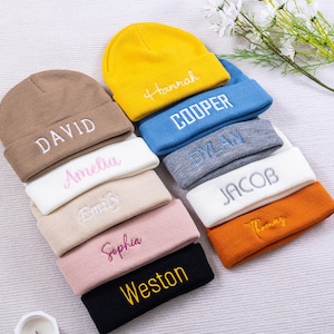 May include: A selection of colourful beanies, each embroidered with a name. The beanies are in shades of brown, yellow, blue, and black, with names like "DAVID", "Hannah", and "COOPER" embroidered in contrasting colours. The beanies are neatly stacked.