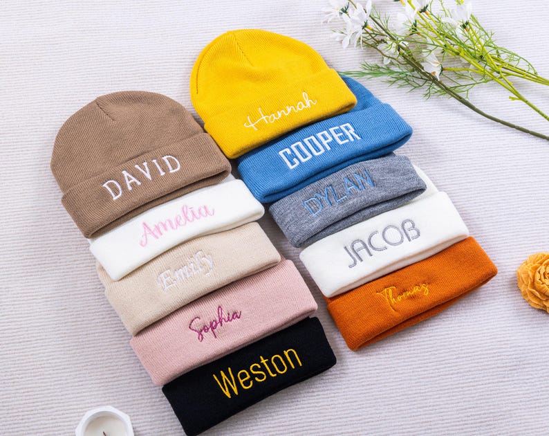 May include: A collection of personalized beanies in various colors, including brown, yellow, blue, and black. Each beanie features an embroidered name in a contrasting color, such as "DAVID", "Hannah", and "COOPER". The beanies are arranged in a neat stack.