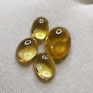 May include: Four oval-shaped, golden-yellow gemstones with a polished, translucent appearance. Each stone has a small, circular, white inclusion. The gemstones are arranged on a light gray, textured surface.