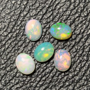 May include: Five oval-shaped opals with iridescent colors of white, green, blue, and yellow. The gemstones are displayed on a textured black surface, showcasing their natural beauty and unique color patterns. These opals are ideal for jewelry making.