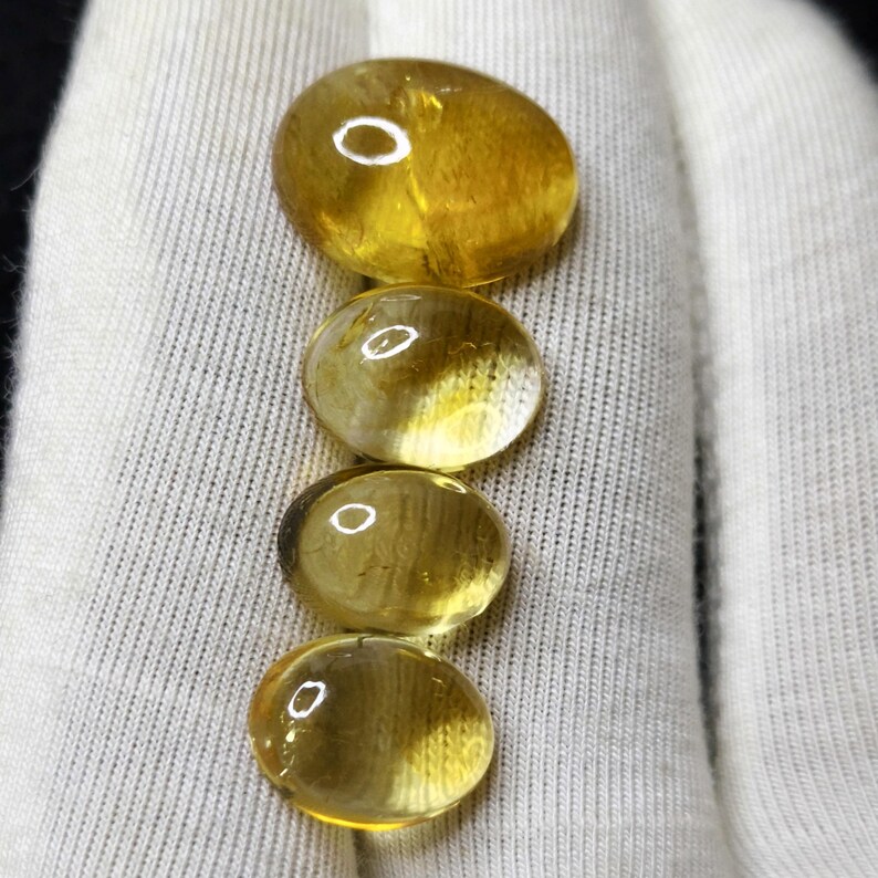 May include: Four oval-shaped, translucent gemstones in shades of yellow and amber are arranged vertically. The stones have a polished, glossy appearance, reflecting light. The background is a white fabric, providing contrast to the gems.