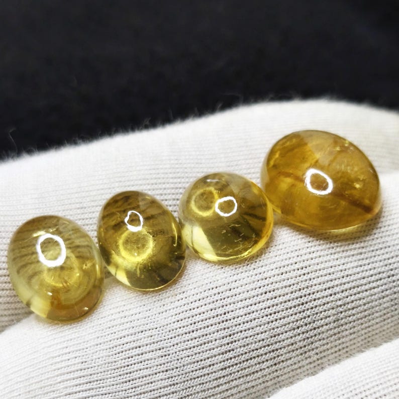 May include: Four oval-shaped, translucent amber gemstones with internal striations and a polished finish. The stones are a warm, golden-yellow color and are displayed on a white fabric surface.