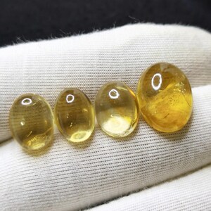 May include: Four polished, oval-shaped gemstones in varying sizes. The stones are a translucent golden-yellow color, with a smooth, glossy surface. They are arranged in a row on a white fabric background, showcasing their natural beauty and clarity.