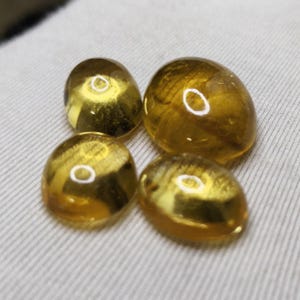 May include: Four polished, oval-shaped gemstones with a golden-yellow hue. The stones are arranged in a cluster, each displaying a smooth, rounded surface. The gems are set against a neutral, off-white fabric background, highlighting their color and clarity.