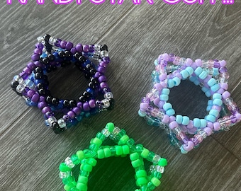May include: Three star-shaped kandi cuffs in the image. One is purple, black, and blue, another is light blue and lavender, and the third is bright green. The text "KANDI STAR CUFF!!!" is above the cuffs.