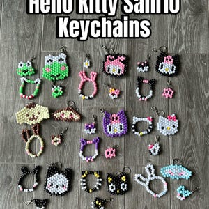 Könnte beinhalten: Eine Sammlung von Hello Kitty Sanrio Schlüsselanhängern in verschiedenen Designs, darunter Hello Kitty, Frösche und Hasen. Die Schlüsselanhänger bestehen aus bunten Perlen und haben silberne Schlüsselringe. Der Text "Hello Kitty Sanrio Keychains" steht oben.