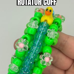 May include: A handmade Kandi cuff bracelet with the text "KANDI DUCK ROTATOR CUFF". The bracelet features bright green beads, clear flower beads with pink centres, and a yellow duck charm. A central column of blue beads adds contrast.