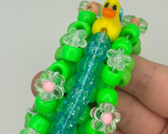 Könnte beinhalten: Ein handgefertigtes Kandi-Armband mit dem Text "KANDI DUCK ROTATOR CUFF". Das Armband hat leuchtend grüne Perlen, transparente Blumenperlen mit rosa Zentren und einen gelben Entenanhänger. Eine zentrale Säule aus blauen Perlen sorgt für Kontrast.