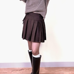 May include: A brown pleated skirt with a matching tie-waist detail. The skirt is paired with a gray sweater, black knee-high boots, and white socks. The outfit is set against a white background.