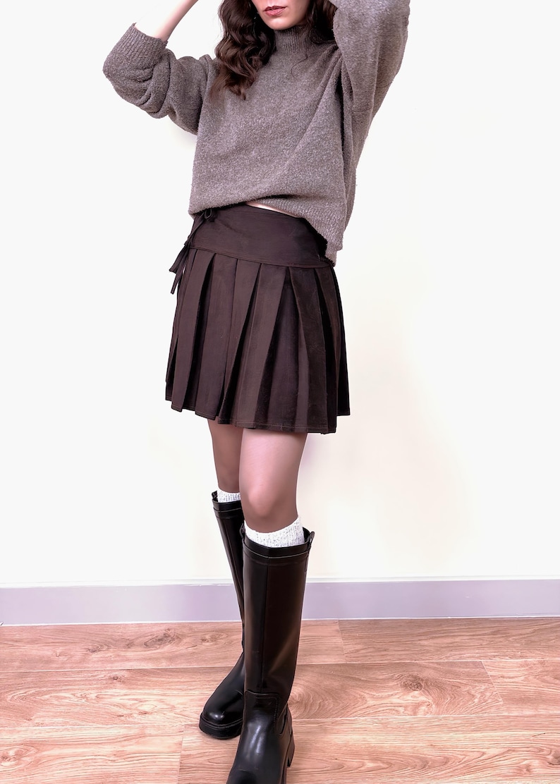 May include: A brown pleated skirt with a matching tie-waist detail. The skirt is paired with a gray sweater, black knee-high boots, and white socks. The outfit is set against a white background.