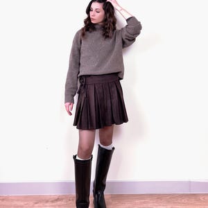 May include: A woman's outfit featuring a brown sweater and a pleated brown skirt. The outfit is completed with black knee-high boots. The sweater has a high neckline. The skirt is tied at the waist. The boots have a glossy finish.