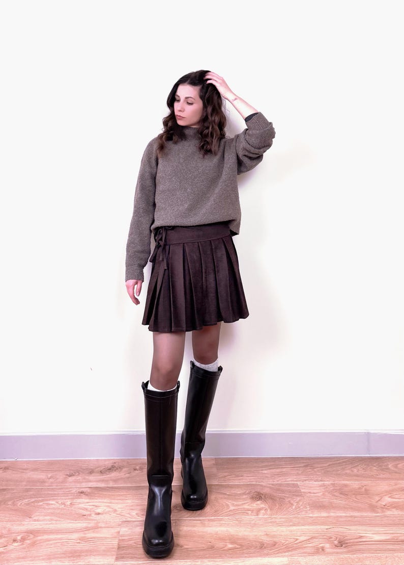 May include: A woman's outfit featuring a brown sweater and a pleated brown skirt. The outfit is completed with black knee-high boots. The sweater has a high neckline. The skirt is tied at the waist. The boots have a glossy finish.