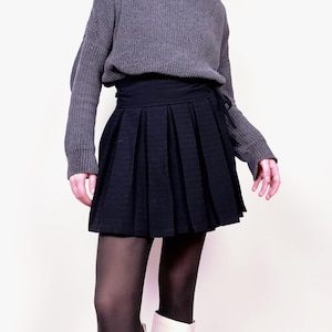 May include: A gray turtleneck sweater paired with a black pleated skirt. The skirt has a wrap-around design. The outfit is completed with black tights and white boots. The overall style is casual and chic.
