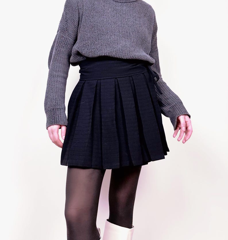 May include: A gray turtleneck sweater paired with a black pleated skirt. The skirt has a wrap-around design. The outfit is completed with black tights and white boots. The overall style is casual and chic.