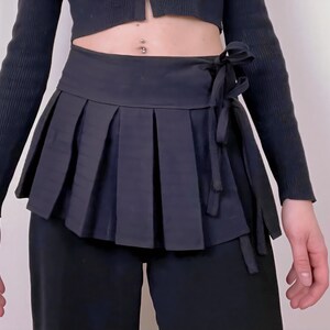 May include: A black pleated wrap skirt with a tie closure. The skirt has a structured, layered design and is worn over black pants. The outfit is completed with a black long-sleeved top.
