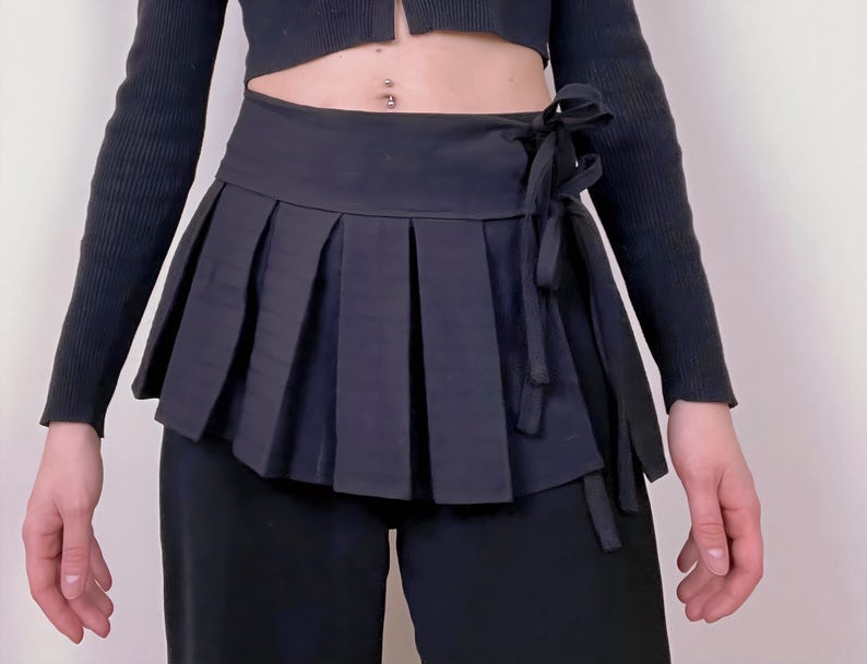 May include: A black pleated wrap skirt with a tie closure. The skirt has a structured, layered design and is worn over black pants. The outfit is completed with a black long-sleeved top.
