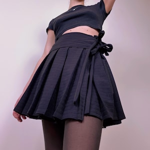 May include: A black pleated mini skirt with a wrap-around tie closure. The skirt has vertical striped detailing and a flared silhouette. The outfit includes a black crop top.