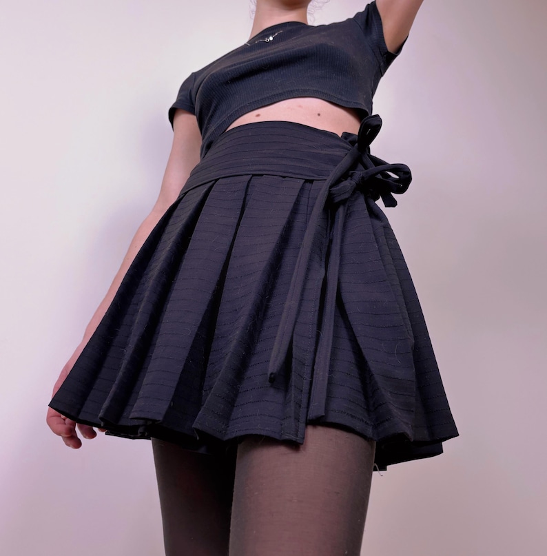 May include: A black pleated mini skirt with a wrap-around tie closure. The skirt has vertical striped detailing and a flared silhouette. The outfit includes a black crop top.