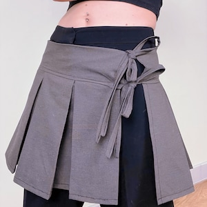 May include: A gray wrap skirt with pleats and a tie closure. The skirt is made of a soft fabric and is worn over black pants. The design features a wrap-around style with a bow detail on the side.