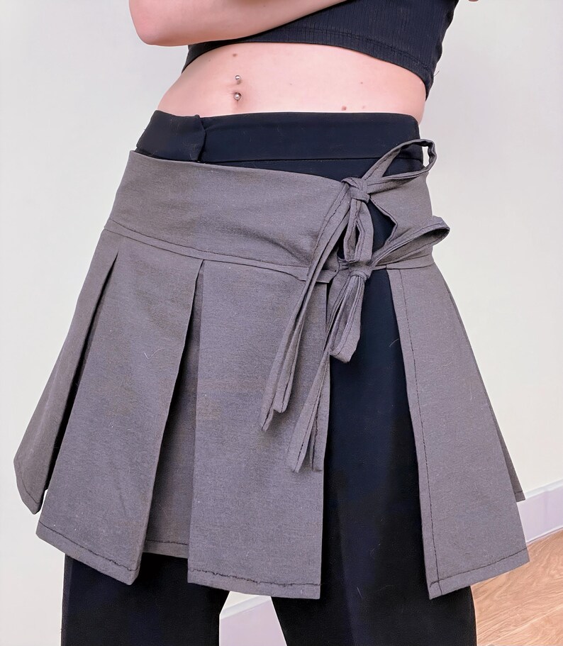 May include: A gray wrap skirt with pleats and a tie closure. The skirt is made of a soft fabric and is worn over black pants. The design features a wrap-around style with a bow detail on the side.