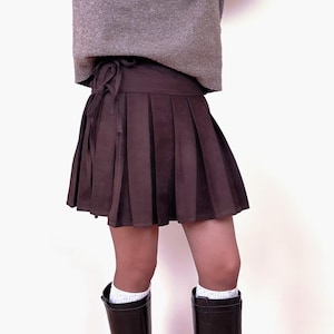 May include: A brown pleated skirt with a tie waist. The skirt is paired with a gray sweater and dark brown boots. White socks are visible above the boots. The skirt is a mini skirt.