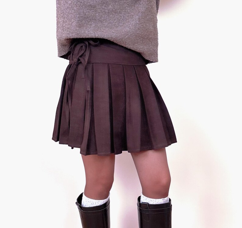 May include: A brown pleated skirt with a tie waist. The skirt is paired with a gray sweater and dark brown boots. White socks are visible above the boots. The skirt is a mini skirt.