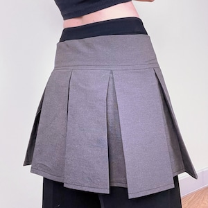 May include: A gray pleated skirt with a high waist. The skirt has a structured design with a flared silhouette. The garment is paired with a black top and black pants. The skirt's fabric appears to be a medium-weight material.