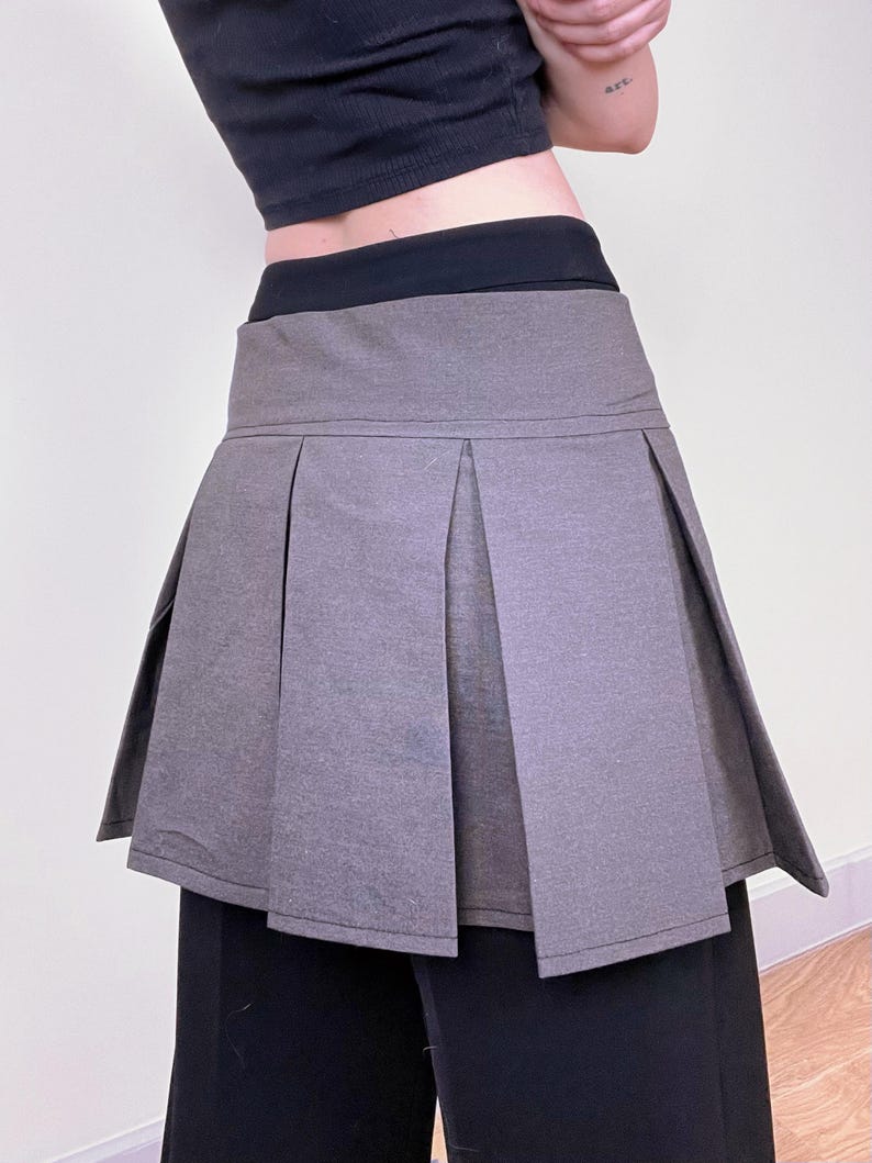May include: A gray pleated skirt with a high waist. The skirt has a structured design with a flared silhouette. The garment is paired with a black top and black pants. The skirt's fabric appears to be a medium-weight material.