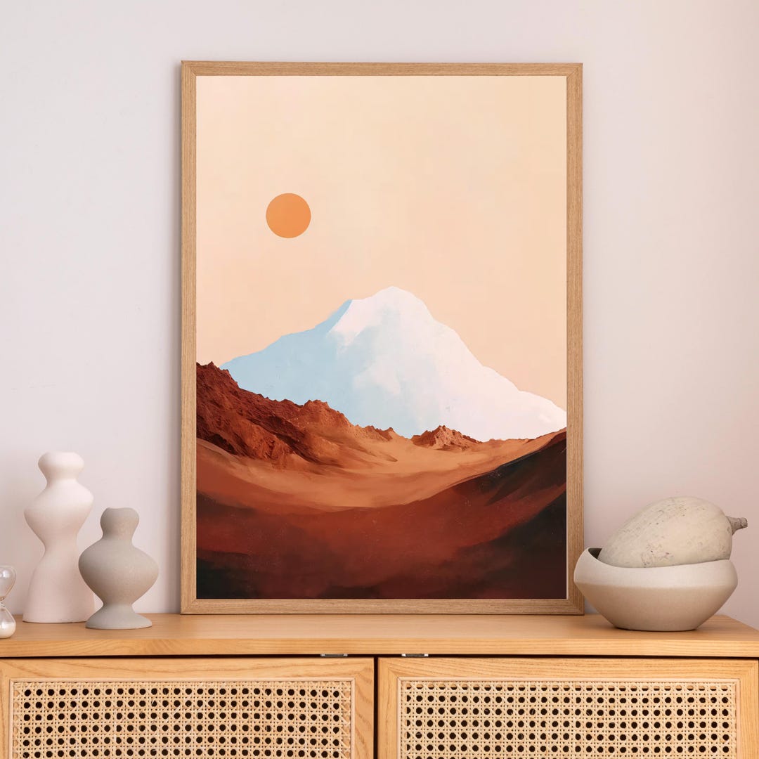 Desert Mountain Wall Art: a snow-capped mountain under a peach sky with a round orange sun, above reddish-brown desert terrain, shown in a light wood frame.
