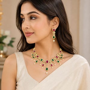 May include: A gold-toned necklace and matching drop earring set featuring alternating rectangular and pear-shaped red and green gemstones. The jewellery is accented with small white stones and tiny dangling white pearls along the lower edge.