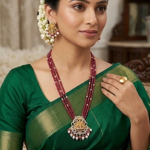 May include: A multi-strand necklace featuring deep red beads and a central gold-toned pendant with intricate detailing, white pearls, and small clear stones. Matching gold-toned drop earrings with similar red beads, white pearls, and clear stones.