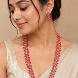 May include: A long gold-tone necklace and matching drop earrings featuring a repeating floral pattern set with vibrant pink stones. The necklace consists of multiple interconnected flower motifs, while each earring displays a single flower design.