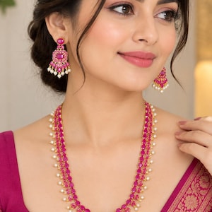 May include: A gold-toned necklace and matching drop earrings set featuring vibrant fuchsia oval-cut stones arranged in a multi-layered design, accented with small white pearls dangling along the edges of the jewelry pieces.