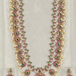 May include: A gold-toned long necklace and matching pair of earrings featuring circular motifs with embedded green and pink stones, clear crystals, and small white pearl-like beads hanging from the edges of the jewelry pieces.
