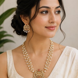 Può includere: Una collana color oro con tre fili di piccole perle bianche e un ciondolo circolare centrale con accenti rossi e verdi e perle pendenti. Orecchini a goccia circolari abbinati con pietre rosse e verdi e piccoli accenti di perle bianche.