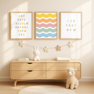 Op de afbeelding: Drie ingelijste prints met houten frames worden tentoongesteld op een lichte houten dressoir. De prints tonen kleurrijke ontwerpen: een alfabet, golvende lijnen en cijfers. Een sterrenslingers, een wit konijnenbeeldje en een schapenknuffel staan ook op de dressoir.