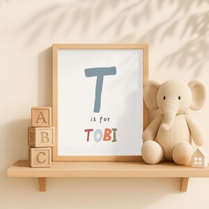 Op de afbeelding: Een ingelijste print met een grote blauwe letter T, de tekst "is for" en de naam "TOBI" in veelkleurige letters. Naast de lijst staan drie gestapelde houten alfabetblokken met A, B en C, en een beige pluche olifant.