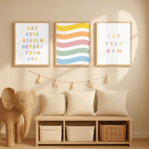 Op de afbeelding: Drie ingelijste prints met houten frames. Eén toont het alfabet in kleurrijke letters, een andere heeft golvende strepen en de derde toont de nummers 1-10. Daaronder een bank met kussens, manden en een rieten olifant.