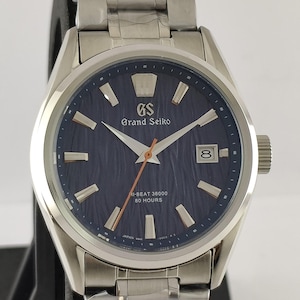 May include: A silver-tone stainless steel wristwatch with a dark blue textured dial, featuring the Grand Seiko logo, a date window at the three o'clock position, and an orange second hand. The watch includes a metal link bracelet with protective film.