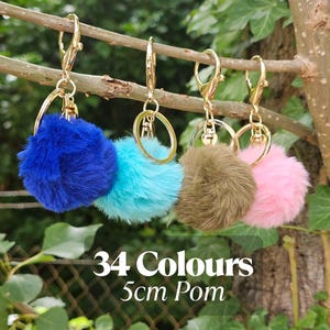 Op de afbeelding: Vier pluizige pompom-sleutelhangers in blauw, turquoise, olijfgroen en roze, elk met een goudkleurige sluiting. De tekst "34 Colours 5cm Pom" staat eronder.
