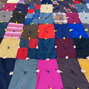 May include: A handmade patchwork quilt constructed from rectangular fabric pieces in various colors and textures, including red, blue, yellow, pink, and plaid. Each fabric square is tied at the center with short lengths of yarn.