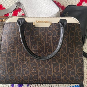 May include: Brown and tan Calvin Klein handbag with black handles. The bag features the Calvin Klein logo in gold and a repeating CK pattern. A white and gray bag and a black bag are visible in the background.