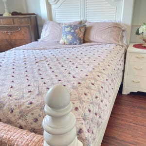 May include: A white bed frame with a headboard and footboard. The bed is made with a floral patterned quilt and matching pillows. A decorative white post is visible at the foot of the bed. The room has a vintage aesthetic.